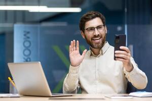 A young businessman man is working in the office on a laptop, sitting at a desk and talking on a call through a phone camera, greeting and waving. photo