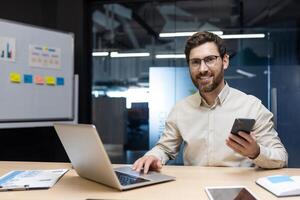 Portrait of a young businessman working in the office, sitting at a desk with a laptop, a board with stickers, holding a phone in his hand and smiling at the camera. photo