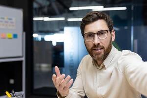 Close-up photo of a young man who is in the office at the workplace, holding a camera in his hand and talking on a call, gesturing with his hand.