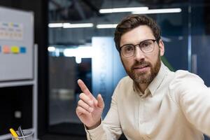 Close-up portrait of a young businessman in the office, pointing to an empty place, talking to the camera. photo