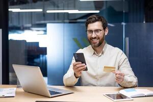 A smiling young man works in the office, sits at a desk, holds a credit card and looks at a mobile phone. photo