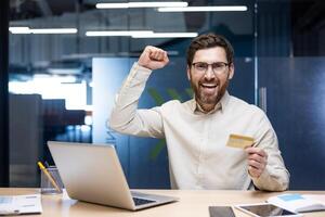 Portrait of a happy young man sitting in the office at a desk with a laptop, holding a credit card in his hands and rejoicing in financial success at the camera, raising his hand up. photo
