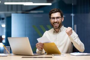 Close-up portrait of a happy young man sitting at a desk in the office. holds an open envelope in his hands, rejoices at the received letter and news. photo