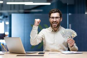 Portrait of a young happy businessman man sitting in the office at the table, rejoicing in financial success, holding a fan of dollar bills in his hand and smiling at the camera. photo