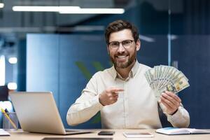 Portrait of a smiling young man sitting in an office at a desk with a laptop, holding a fan of banknotes and pointing at dollars. photo