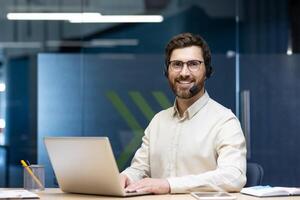 Portrait of a smiling young man in a headset sitting at a desk in the office, using a laptop and looking confidently at the camera. photo