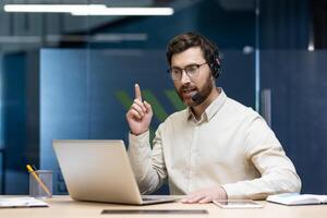 Serious young man sitting in the office at the desk in a headset and talking on a call on a laptop, explaining, pointing. photo