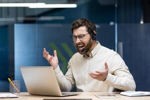 Young angry man in headset working in office on notebook, sitting at desk and emotionally shouting and arguing. photo