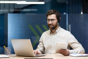 A young man in a headset is sitting at a desk in the office, holding a notebook and working on a laptop. photo