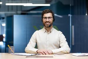 Portrait of a young man in a headset sitting in front of the camera in the office at the table and smilingly looking ahead. photo