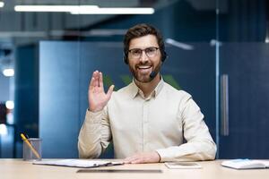 Portrait of a happy and successful young man in a headset sitting at a table in the office in front of the camera, smiling online, greeting and waving. photo