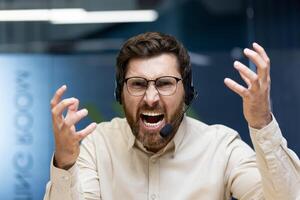 Portrait of a young angry man in a headset sitting in the office in front of the camera and angrily talking on a call while waving his hands. photo