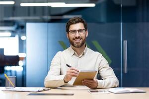 Portrait of a smiling young man sitting in the office at the desk in front of the camera, holding a notebook and a pen in his hands. photo