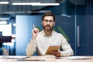 A young male office worker sits at a desk in front of the camera, holds a notebook in his hand and points his finger up to an empty place. photo