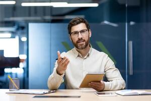 A young man is sitting in the office at the table, holding a notebook with a pen in his hands, talking to the camera on a call, gesturing with his hands. photo
