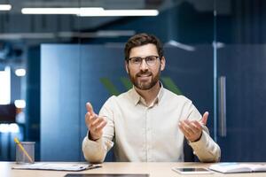 Portrait of a young man sitting in the office at a table in front of the camera and talking on a call, gesturing to the camera. photo