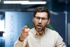Close-up photo of a young angry businessman sitting in an office with a desk in front of the camera and talking angrily on a call while pointing his finger at the camera.