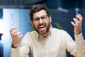 Close-up portrait of a young man in glasses and a shirt who is in the office and angrily talking and shouting at the camera while gesturing with his hands. photo