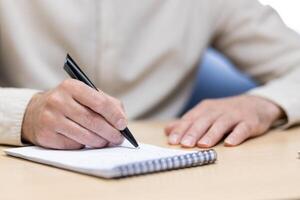 Close-up photo of a part of the body, hands of a young man in a light shirt holding a pen and making notes in a notebook at a desk in the office.