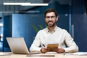 Portrait of a young man in glasses sitting in the office at a desk with a laptop, holding a notebook and looking at the camera with a smile. photo