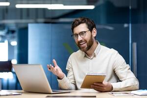 A smiling young man works in the office, sits at a workplace in front of a laptop, holds a notebook with a pen in his hands, conducts a meeting. photo