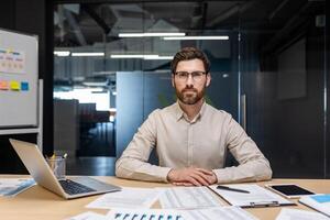 Portrait of a young serious and self-confident man sitting in the office at a desk behind a laptop and working with a project and documents. photo