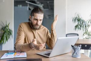 A man in a brown shirt, looks at a laptop with a confused expression, gesturing with his hands as if in disbelief. photo