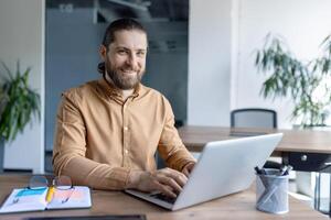A bearded man smiles at the camera while working on a laptop. The office environment shows the focus on productivity and work. photo