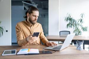 Bearded man works at a laptop while also using a mobile phone at his desk in a brightly lit modern office, looking focused. photo