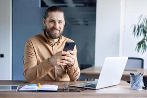 A smiling man with a beard uses his smartphone while sitting at his desk, focused and engaged. The open laptop hints at work. photo