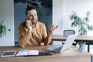 A smiling businessman is on a phone call, gesturing with his hand. He is seated at a desk with a laptop and other office supplies visible. photo