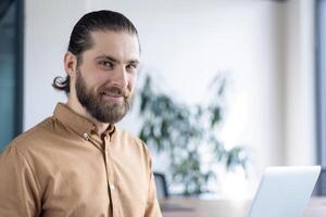 A smiling businessman with a beard looks towards the camera in an office setting. He is wearing a brown shirt and sitting near a laptop on a desk. photo