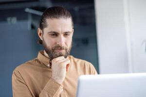 A bearded man with a ponytail looks down at a laptop, pondering a thought while working in an office setting. photo