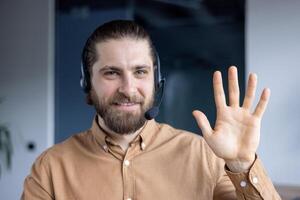 A smiling man with a headset waves at the camera, friendly and inviting. He is wearing a button-down shirt. photo