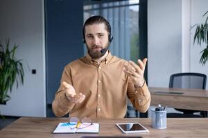 A bearded man wearing a headset gestures with both hands while sitting at a desk, ready for a call or online meeting. photo