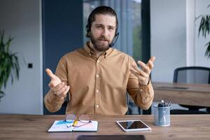 A bearded man wearing a headset gestures while sitting at his desk with a tablet, notebook, and glasses, during a virtual meeting. photo