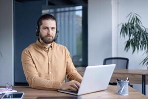 A man with a headset working on his laptop in the office he is ready to assist clients with a friendly, professional demeanor. photo