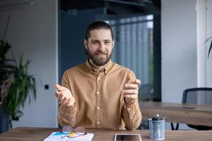 A man with a beard gestures during a call, seated at a wooden desk in a modern office environment. He appears engaged and approachable. photo