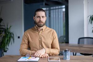 A man with a beard sits at his desk with a tablet and notebook, in a modern office setting. He has a serious look and is staring ahead. photo