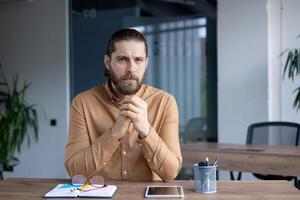 A thoughtful man with a beard, looking directly at the camera. He is sitting at a wooden desk with a notepad and glasses in front of him. photo