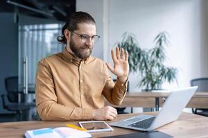 A bearded man waves during a call, smiling at the laptop in front of him in a modern office setting. photo
