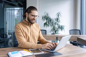 A businessman with glasses is working on his laptop at a wooden desk in an office setting with a plant and notebook in view. photo