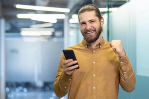 A man celebrates success while holding a smartphone in an office setting. The man's joyful expression and gesture capture a moment of achievement. photo