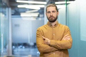 A confident man with a beard and arms crossed stands in a modern office setting, looking directly at the camera. photo