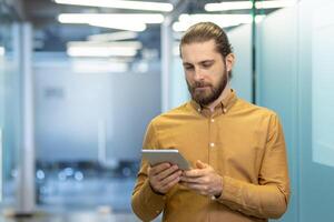 Man with a beard in an office using a tablet to communicate. He is wearing a mustard-colored shirt while looking at the screen. photo