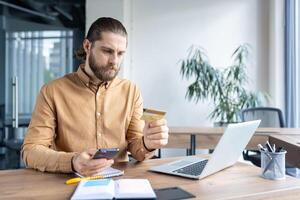 A focused man is holding a credit card and smartphone while working at a desk with a laptop. He is looking intently at the screen. photo