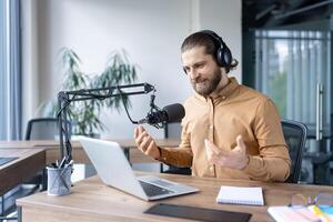 A man with a beard uses a microphone and laptop, engaging in a live podcast recording in an office setting. photo