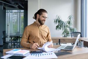 Man with glasses reviews documents at an office desk with a laptop, calculator, and graphs, focused on financial analysis and planning. photo