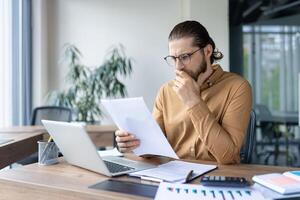 A focused professional reviews documents at his desk in an office setting, pondering the information. Laptop, papers, and calculator are visible. photo