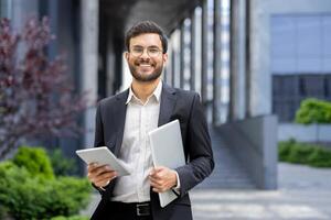 A smiling businessman holds a tablet and a laptop, showing his modern, digital tools. The man is well-dressed with glasses in front of a modern building. photo